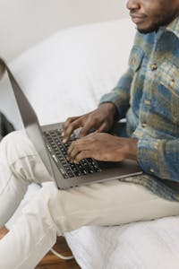 From above of crop concentrated African American male freelancer in casual clothes working remotely on laptop while sitting on comfortable bed at home